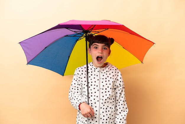 Menina segurando um guarda-chuva isolado na parede bege com expressão facial surpresa