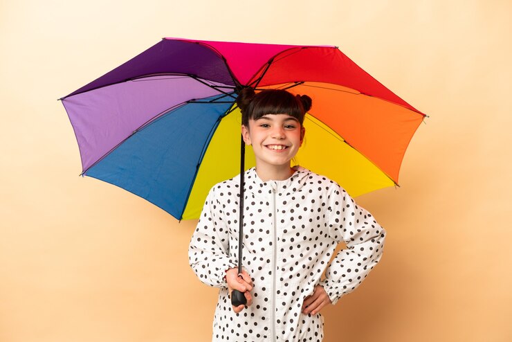 Foto menina segurando um guarda-chuva isolado na parede bege, posando com os braços na cintura e sorrindo