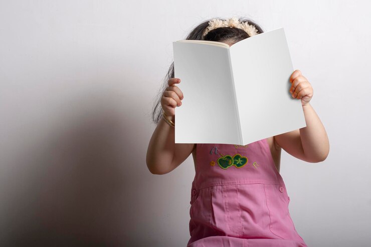 Foto menina segurando um livro de histórias infantis com capa em branco na frente de seu rosto, modelo de simulação editável pronto para seu design, caminho de seleção de capa incluído