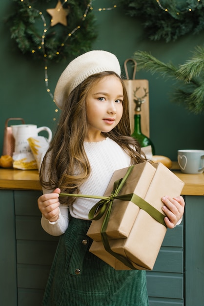 Menina segurando um presente de natal em uma boina bege, blusa bege e saia turquesa. desembalando um presente de natal