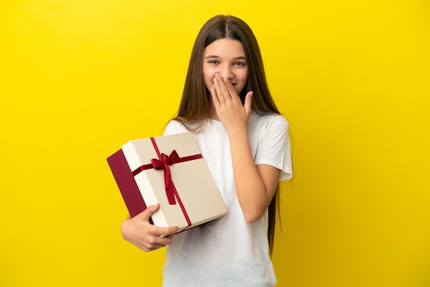 Menina segurando um presente sobre um fundo amarelo isolado, feliz e sorridente, cobrindo a boca com a mão