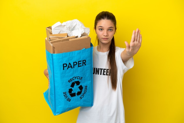Menina segurando um saco de reciclagem cheio de papel para reciclar sobre um fundo amarelo isolado fazendo um gesto de pare