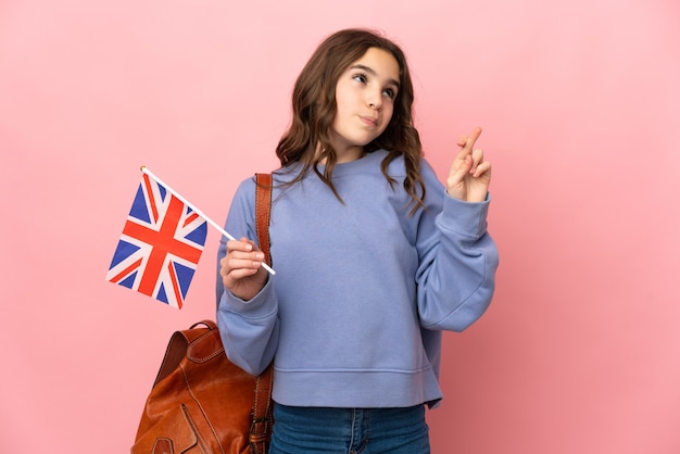 Menina segurando uma bandeira do Reino Unido isolada em um fundo rosa, cruzando os dedos e desejando o melhor