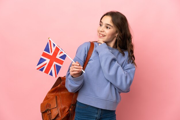 Menina segurando uma bandeira do Reino Unido isolada em um fundo rosa, olhando para cima enquanto sorri