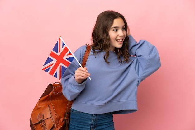 Menina segurando uma bandeira do Reino Unido isolada em um fundo rosa, ouvindo algo colocando a mão na orelha