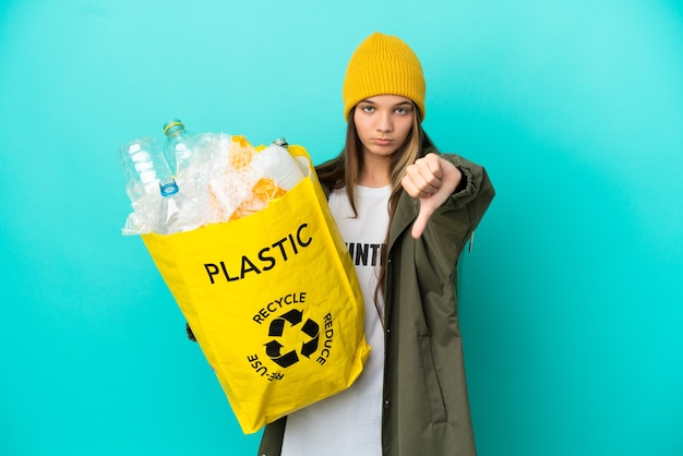 Menina segurando uma sacola cheia de garrafas plásticas para reciclar sobre fundo azul isolado, mostrando o polegar para baixo com expressão negativa