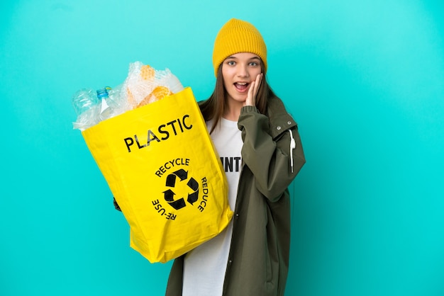 Menina segurando uma sacola cheia de garrafas plásticas para reciclar sobre um fundo azul isolado com surpresa e expressão facial chocada