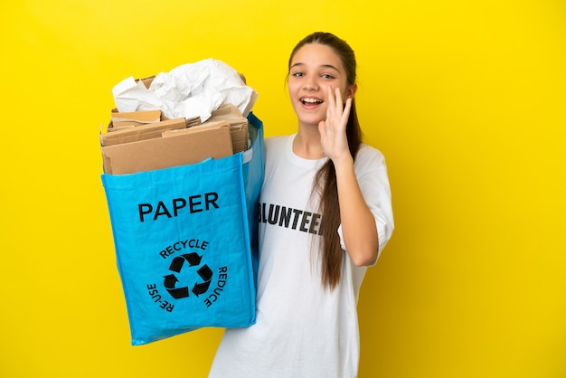 Menina segurando uma sacola cheia de papel para reciclar sobre uma superfície amarela isolada, gritando com a boca aberta