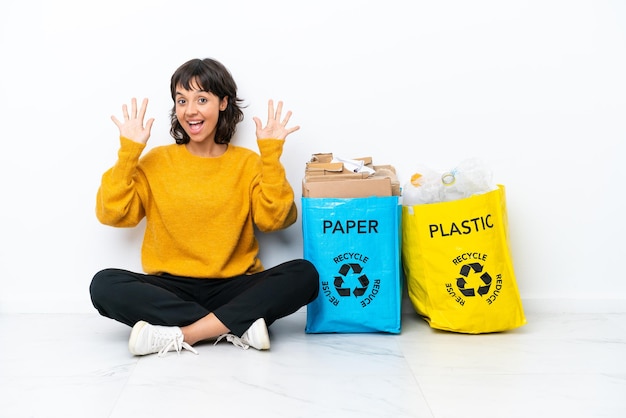 Foto menina segurando uma sacola cheia de plástico e papel, sentada no chão, isolada no fundo branco, contando dez com os dedos