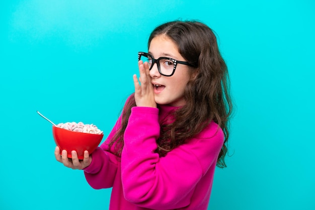 Menina segurando uma tigela de cereais isolada em fundo azul sussurrando algo