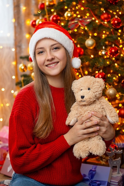 Menina segurando ursinho de pelúcia perto de árvore de Natal decorada Celebração de Natal em casa decorações festivas ao redor