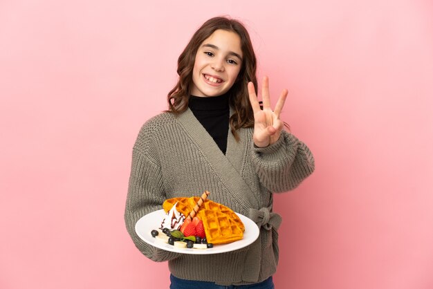 Menina segurando waffles isolados em um fundo rosa feliz e contando três com os dedos