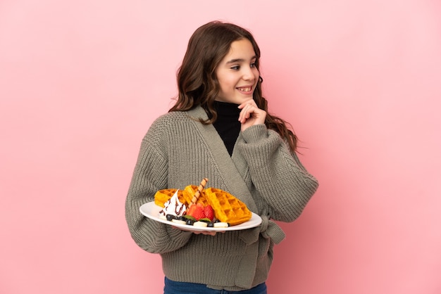 Menina segurando waffles isolados em um fundo rosa, olhando para o lado e sorrindo