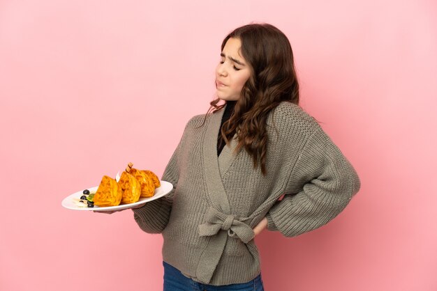 Foto menina segurando waffles isolados em um fundo rosa, sofrendo de dor nas costas por ter feito um esforço