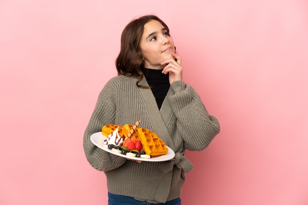 Menina segurando waffles isolados em um fundo rosa tendo dúvidas enquanto olha para cima