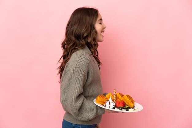 Menina segurando waffles isolados na parede rosa rindo na posição lateral