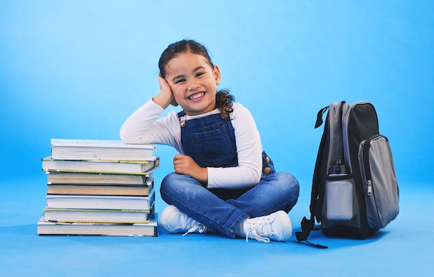 Menina sentada e livros em mochila de retrato de estúdio e animada para aprender no chão por fundo azul Educação e desenvolvimento de criança feminina com sorriso futuro e estudando para conhecimento