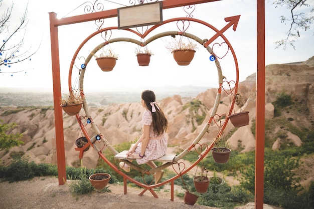 Foto menina sentada em um banco decorado em forma de coração em um ponto de vista e admirando a vista da capadocia