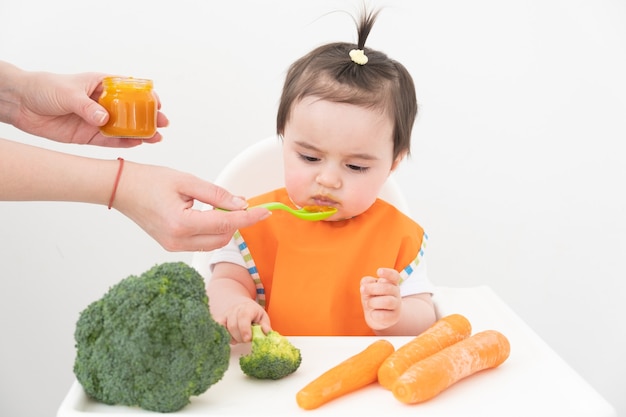 Menina sentada em uma cadeira de childs comendo purê de vegetais. mamãe alimenta o bebê.