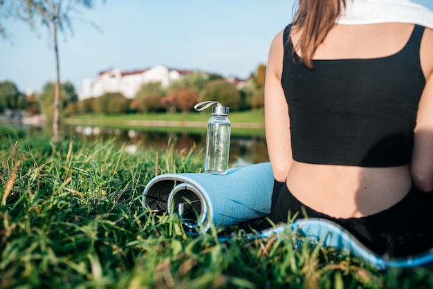 Menina sentada na grama com garrafa e tapete de ioga. Fitness na cidade metrópole na grama no verão ao ar livre