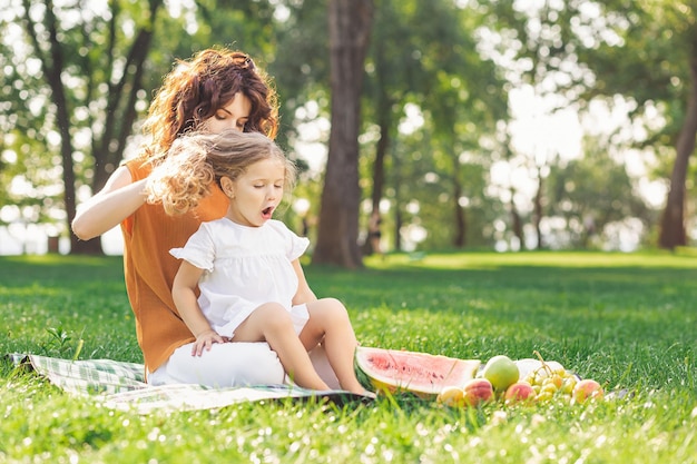 Menina sentada nas pernas da mãe enquanto ela faz o cabelo no parque