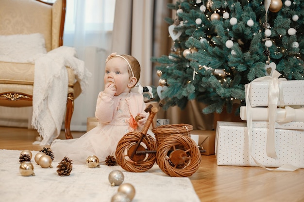 Menina sentada no chão perto da árvore de natal em casa. Garota está brincando com bolas para decoração de árvore de Natal. Menina com vestido pêssego.