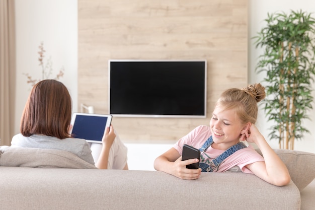 Menina sentada no sofá e assistindo algo interessante no celular enquanto a mãe assistindo tv, conceito de surpresa