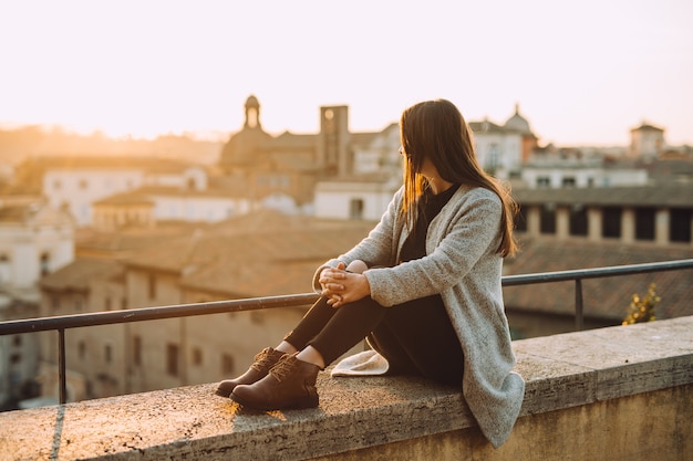 Menina sentada no topo do prédio com vista para a cidade e um lindo pôr do sol