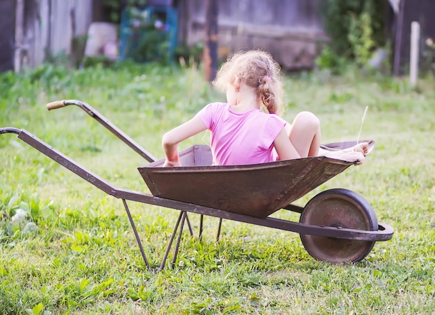 Menina sentada no velho carrinho de mão de ferro na aldeia no verão