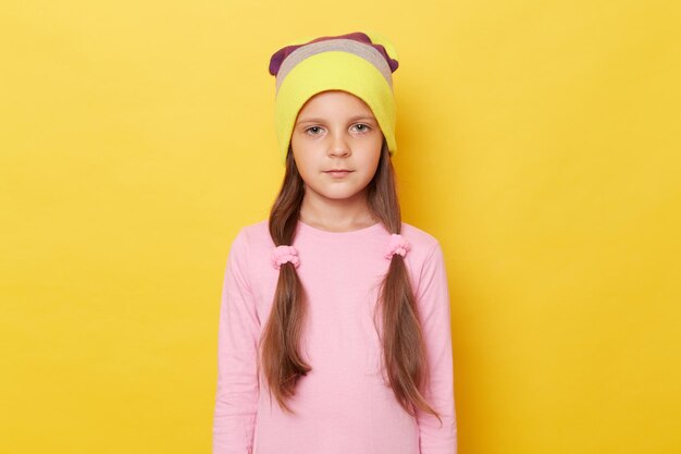 Menina séria e confiante com rabo de cavalo vestindo camisa rosa e gorro posando isolada sobre fundo amarelo olhando para a câmera com expressão descontente