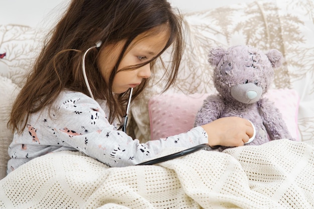 Menina séria em pijamas sentada debaixo do cobertor e ouvindo o pulmão do ursinho de pelúcia com estetoscópio Repouso na cama