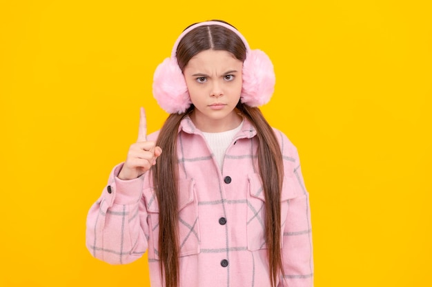 Menina séria em protetores de ouvido e sobretudo de inverno mantém o dedo apontando para cima a ideia de fundo amarelo