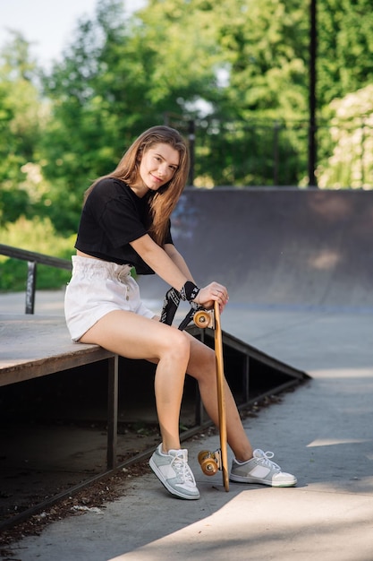 Menina skatista adolescente com um skate sentado no parque de skatistas