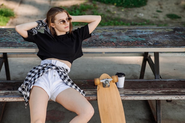 Menina skatista adolescente eith um skate está relaxando no banco no parque de skatistas