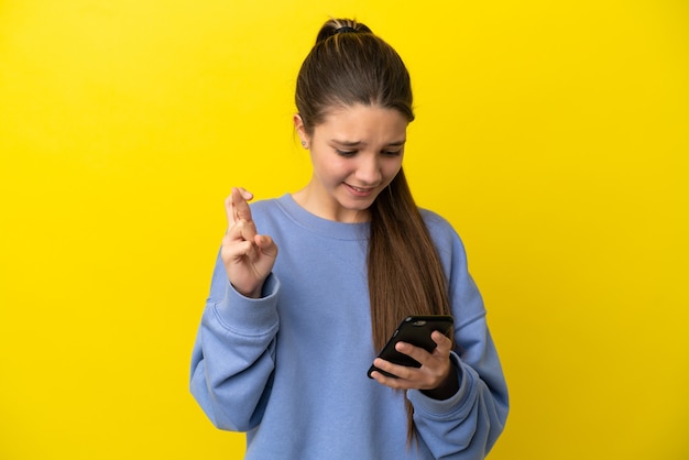 Menina sobre fundo amarelo isolado usando telefone celular com os dedos se cruzando