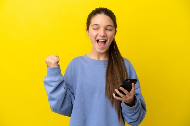 Menina sobre fundo amarelo isolado usando telefone celular e fazendo gesto de vitória