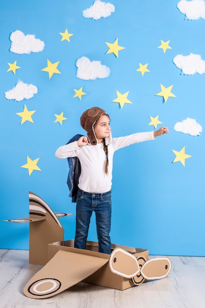 Menina sonhadora brincando com um avião de papelão no estúdio com fundo de céu azul e nuvens brancas.