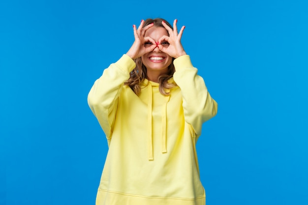 Menina sonhadora e boba, olhando por entre os dedos com uma expressão divertida e feliz, faça gestos bem formando óculos com as mãos e sorrindo despreocupada, pareça distante, procurando algo, em uma parede azul