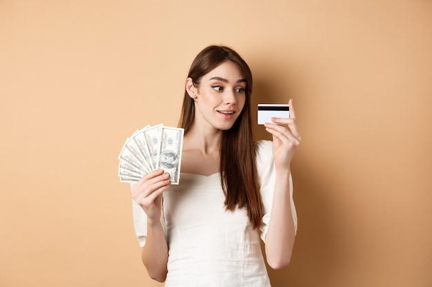 Menina sonhadora, olhando para o cartão de crédito de plástico e pensando em fazer compras, segurando notas de dólar, de pé sobre fundo bege.