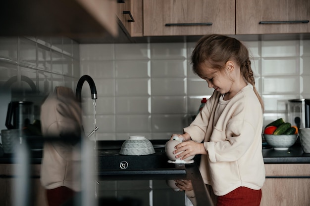 Menina sorridente ajuda nas tarefas domésticas lava pratos na cozinha