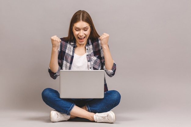 Menina sorridente animada, sentada no chão com o laptop, levantando uma mão no ar é que ela ganha