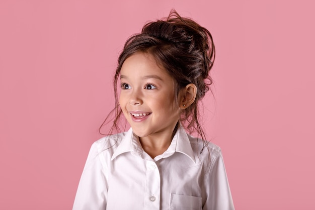 Menina sorridente bonitinha na camisa branca com penteado, olhando para a câmera no fundo rosa. Emoções humanas e expressão facial