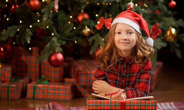Menina sorridente com chapéu de Papai Noel, sentada perto da árvore de Natal e escrevendo uma carta para o Papai Noel.