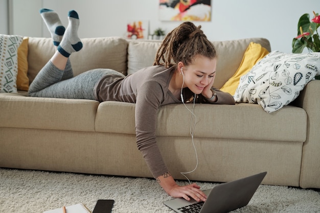 Menina sorridente com fones de ouvido deitada em um sofá confortável e assistindo a um vídeo no display do laptop no chão de casa