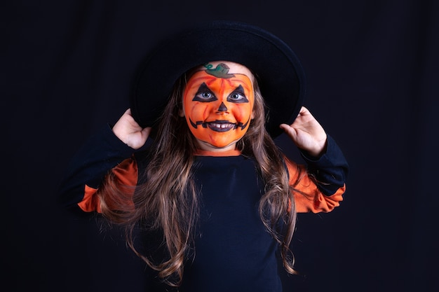 Menina sorridente com maquiagem de abóbora no rosto, segurando um chapéu preto em uma parede preta, festa de halloween engraçada.