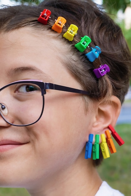 Foto menina sorridente com óculos com presilhas coloridas de arco-íris e brincos coloridos com prendedores de roupa nas orelhas, close-up, tema lgbtq