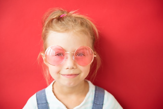 Menina sorridente com óculos grandes redondos cor de rosa sobre fundo vermelho