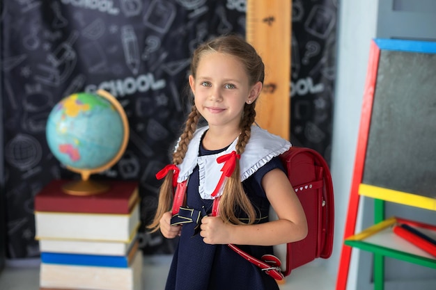 Menina sorridente com tranças e em um uniforme escolar, costas vermelhas fica em sala de aula na escola.