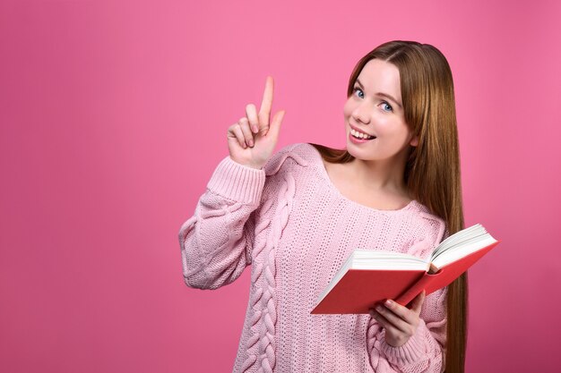 Menina sorridente com um livro na mão aponta para cima com o dedo indicador.