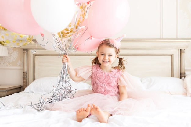 Menina sorridente com vestido sentado na cama com saltos nus e segurando pacotes de balões de aniversário infantil